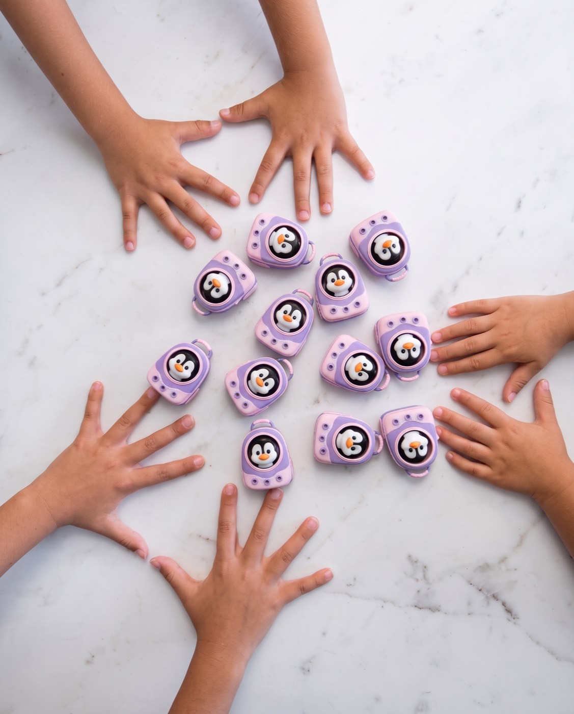 Six children's hands reaching in around a cluster of purple 3D-printed penguins on a marble surface.